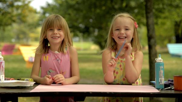 Zwei Mädchen sitzen an einem Tisch und lachen in die Kamera. Vor ihnen liegt ein rosanes Papier.