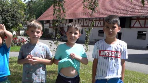 Vier Jungen stehen auf einer Wiese vor einem Bauernhofgebäude. Sie tragen T-Shirts und kurze Hosen.