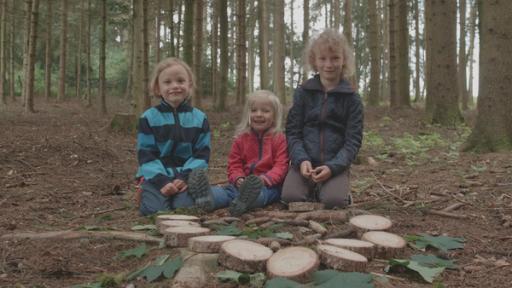 Drei Kinder sitzen auf dem Waldboden. Vor ihnen liegen verschiedene Naturmaterialien.