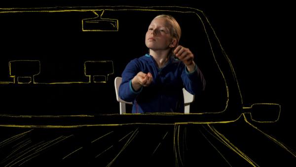 Ein Mädchen sitzt im Auto und bewegt ihre Hände so, als würde sie ein Lenkrad halten. 
