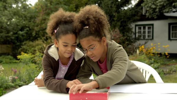 Zwei Mädchen sitzen draußen im Garten und suchen sich Stifte aus einer Box heraus. 