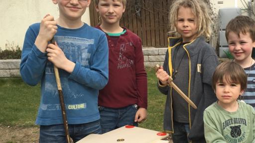 Kinder stehen an einer Werkbank. Vor ihnen liegt Holz, Schrauben und Werkzeug.