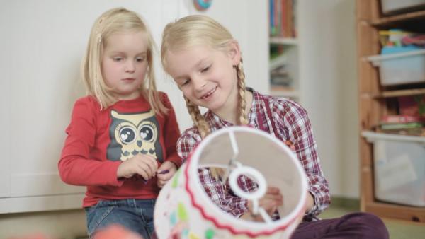 Zwei Mädchen sitzen auf dem Boden und betrachten einen bunt verzierten Lampenschirm.