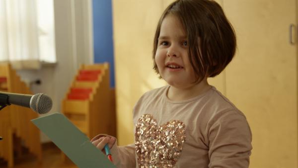 Ein Mädchen mit braunem Haar steht vor einem Mikrofon und hält dabei einen Zettel in der Hand.