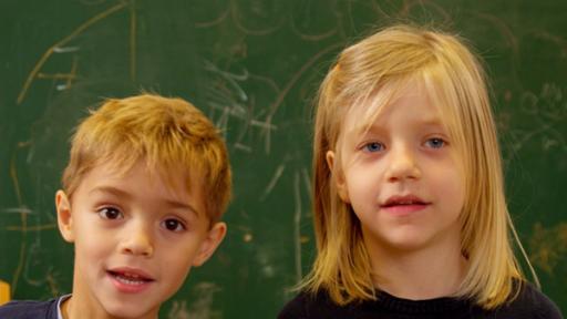 Zwei Kinder stehen vor einer Wand mit einer Tafel und schauen in die Kamera um sich vorzustellen.