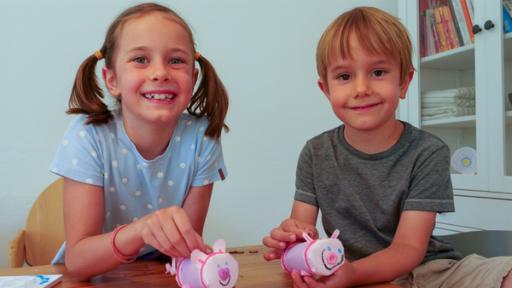 Zwei Kinder sitzen am Tisch und halten rosafarbene Schweine aus Pappe in ihren Händen.