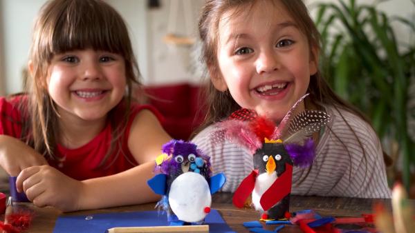 Zwei Mädchen sitzen lachend am Tisch. Vor ihnen stehen zwei bunte Pinguine aus Papier.