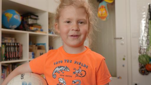 Ein Grundschulkind steht im Kinderzimmer. Es lächelt und hat einen Fußball unterm Arm.