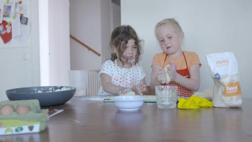 Zwei Mädchen sitzen an einem großen Tisch und kneten Teig. Neben ihnen stehen Materialien wie eine blaue Schüssel und eine Tüte Mehl.