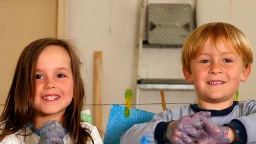 Zwei Vorschulkinder mit Händen voller bunter Farbe stehen vor einer Wäscheleine mit Bildern.