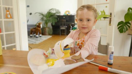 Ein blondes Mädchen zeigt Batelutensilien auf einem Tisch. Darunter ist ein Schwamm, Papiere, ein Klebstift und Malstifte.