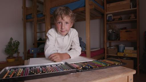 Ein Junge mit Hemd sitzt am Tisch. Vor ihm liegen Buntstifte, die in einer Kiste sortiert sind.