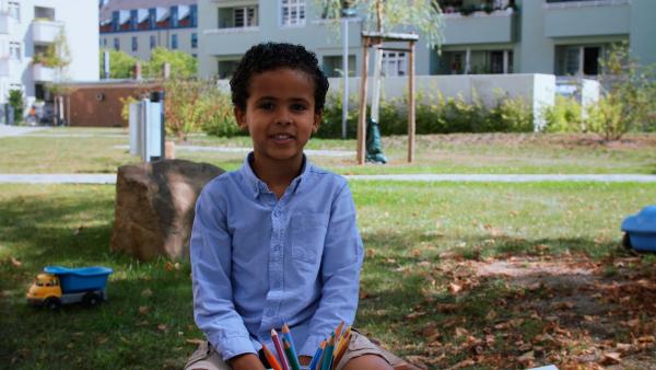 Eine Junge im blauen Hemd sitzt auf einer Wiese und lacht in die Kamera. Vor ihm stehen Buntstifte.