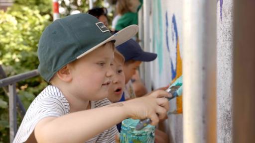 Ein Junge mit einer Kappe steht vor einer Mauer. Er hat ein Glas mit Farbe und einen Pinsel in der Hand. Er streicht die Farbe an die Wand. Im Hintergrund sind weitere Kinder zu sehen.