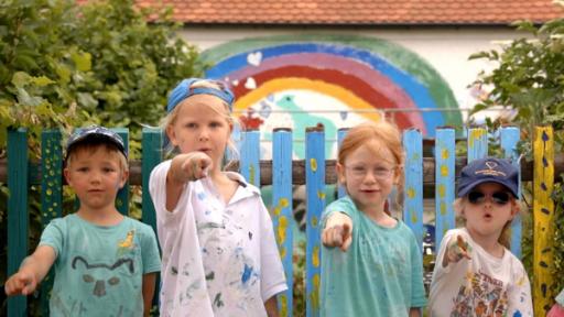 Fünf Kindergartenkinder stehen vor einer bemalten Hauswand. Sie zeigen mit den Fingern nach vorn.