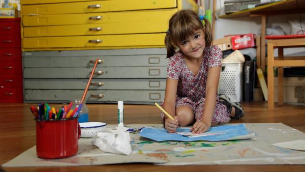 Ein Mädchen kniet auf dem Boden und malt an ihrem Bild. Vor ihr liegen Stifte und Wasserfarben.
