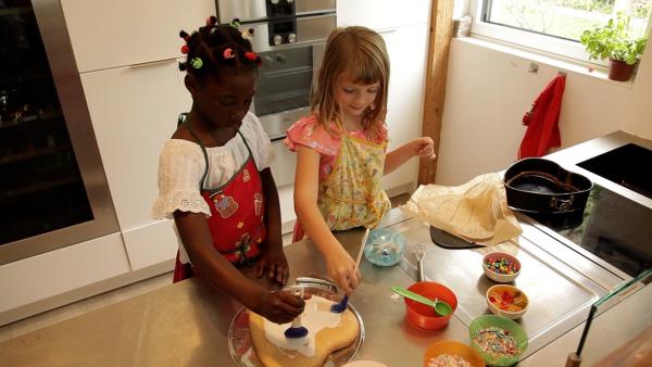 Aminata und Magdalena stehen in der Küche und tragen jeweils eine Schürze. Sie bestreichen einen Kuchen aus Herzform mit Zuckerguss. Auf der Arbeitsfläche steht mehrere kleine Schüsseln mit Süßigkeiten wie Gummibärchen.