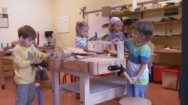 Anastasia, Leopold, Meo und Johannah stehen um eine Werkbank herum. Alle haben Werkzeuge in der Hand und bauen etwas mit Holz.