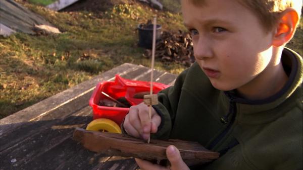 Rechts ein Junge mit einem Holzboot in einer Hand. Mit der anderen Hand hält er einen dünnen Holzstab als Mast für das Boot. Neben ihm ein Tisch mit einer kleinen Werkzeugkiste auf Rädern.