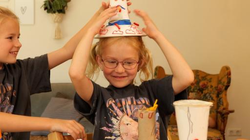 Stella und Stina sitzen an einem Tisch. Vor ihnen liegen verschiedene Bastelmaterialien. Stina hat einen selbst gebastelten und bemalten Hut auf.