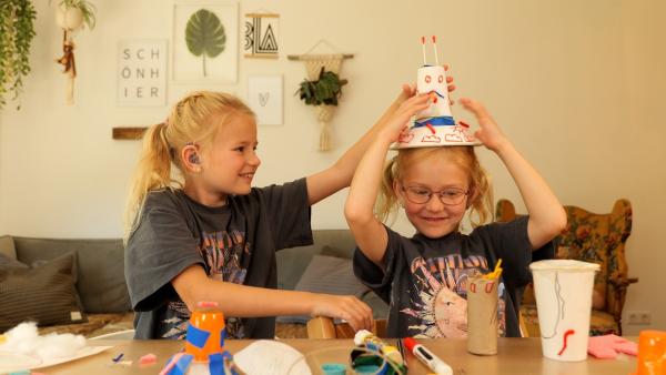 Stella und Stina sitzen an einem Tisch. Vor ihnen liegen verschiedene Bastelmaterialien. Stina hat einen selbst gebastelten und bemalten Hut auf.