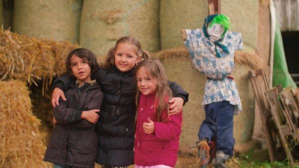 Drei Kinder stehen in Winterjacken vor großen Heuballen. Daneben steht ihre selbst gebaute Vogelscheuche.