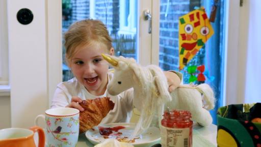 Julia sitzt mit ihrem Kuscheleinhorn am Frühstückstisch und füttert es mit einem Croissant.