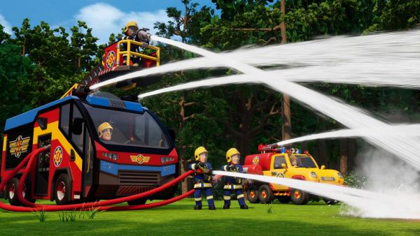 Vor einem Feuerwehrauto stehen zwei Feuerwehrleute und lassen aus dicken Schläuchen Wasser schießen.