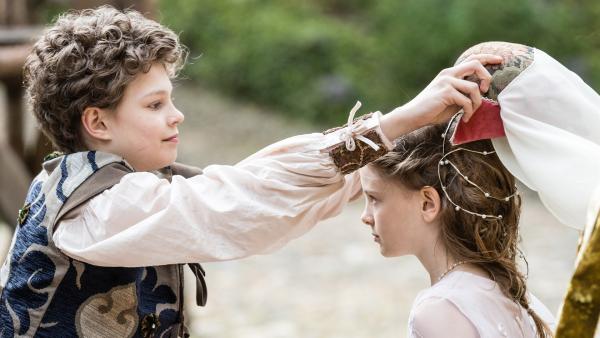 Links steht Leopold seitlich. Mit ausgestreckten Arme hält er einen Haarschmuck mit Schleier über Josefinas Kopf. Josefina schaut zu ihm.