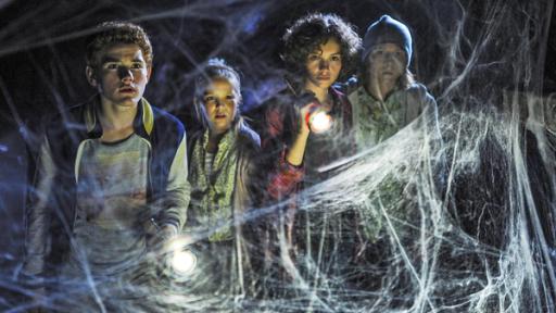 Von links nach rechts: Die "Fünf Freunde" tappen im Dunkeln mit Taschenlampen in der Hand. Julian (Quirin Oettl), Anne (Neele Marie Nickel), Dick (Justus Schlingensiepen) und George (Valeria Eisenbart). Vor ihnen ein dichtes Netz aus Spinnenweben. 