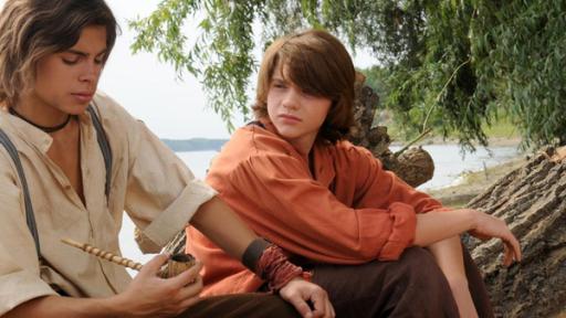 Tom Sawyer (Joel Courtney) und Huckleberry Finn (Jake T. Austin) sitzen am Ufer des Mississippi auf einem Baumstamm, Tom schaut Huck an, dieser ist mit seiner Pfeife beschäftigt.