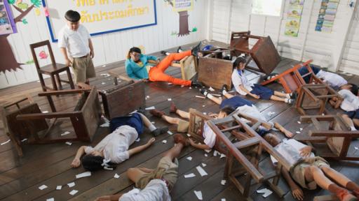 Ein Junge steht in einem Klassenzimmer vor einer Tafel und schaut bedächtig nach unten. Neben ihm liegen Mitschüler bewusstlos am Boden. 