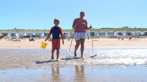 Hugo steht mit seiner Mutter am Strand. Sie sind mit Eimer und Netzen ausgerüstet. Im Hintergrund sind Strandhäuser zu sehen.