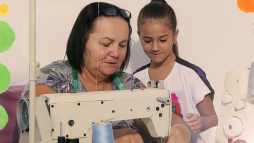 Das Mädchen Maša sitzt neben ihrer Oma, die mit einer Nähmaschine näht. Maša schaut ihr genau zu, denn sie möchte auch Nähen lernen.