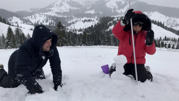 Im Schnee steht Ben links auf allen vieren. Rechts kniet Jess im Schnee und hält einen Zollstock vor sich, mit dem sie etwas vor sich misst. Im Hintergrund sind mit Schnee bedeckte Berge.