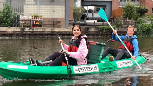 In einem grünen Kajak sitzen Jess (vorne links) und ein Künstler (hintern rechts). Zwischen ihnen steht eineTonne. Auf dem Kajak steht "#Green Kajak".