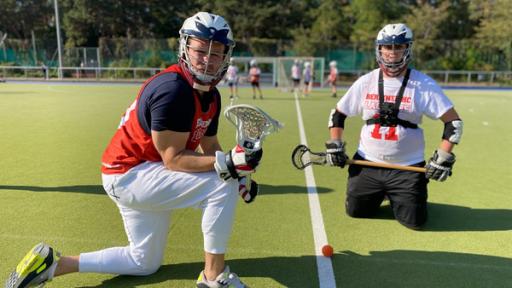 Ben kniet mit Sportklamotten, Helm und Lacrosse Schläger auf einem Spielfeld. Rechts neben ihm kniet ein Lacrosse Spieler.