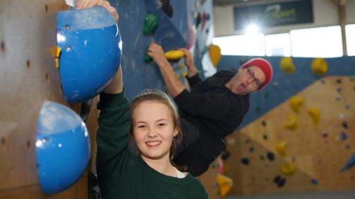 Vorne steht Ana, die sich mit ihrem rechten Arm an einem Griff an einer Kletterwand. Hinter ihr sieht man Ben, der an einer Wand klettert.