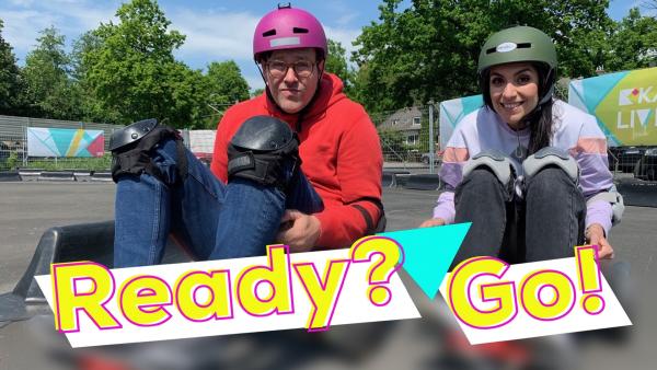 Links Ben und Rechts Jess, die auf verschiedenen Funsport-Geräten sitzen. Sie tragen beide einen Helm und Knie-Schützer. SIe lächeln in die Kamera. Vorne steht die Überschrift auf weißem Banner in gelber Schrift "Ready? Go!".