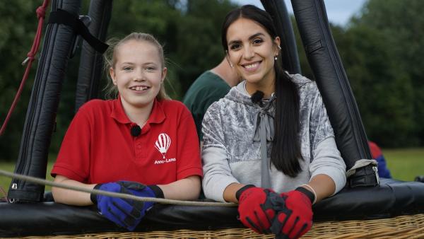 Anna-Lena und Jess stehen im Passagierkorb des Heißluftballons. Sie tragen Schutzhandschuhe und lächeln.