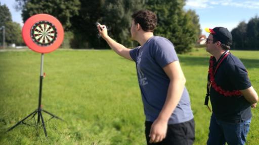 Rechts steht Ben mit einer Deutschland-Kappe und einer Deutschland-Flagge auf der Wange gemalt. Neben ihm steht Dart-Spieler Fabian, in Wurf Position und einem Dartpfeil in der Hand. Links weiter hinten steht eine Dartscheibe.