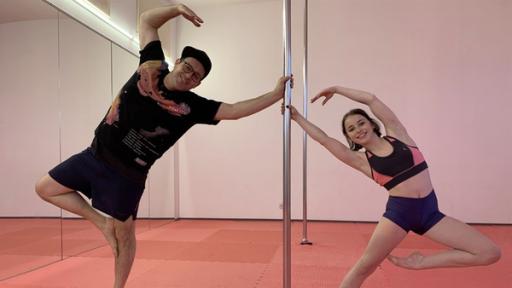 In einem Tanzssal ist in der Mitte eine Poledance-Stange. Links davon Ben, der auf einem Bein steht und sich an der Stange festhält. Rechts ist Finja, die ebenfalls auf einem Bein steht und sich mit einer Hand an der Stange festhält.