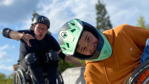 Im Vordergrund Amos im Rollstuhl, der sich mit einem Arm am Boden abstürzt. Er trägt einen Motorrad-Helm. Im Hintergrund sitzt Ben ebenfalls im Rollstuhl.