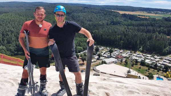 Johannes (links) und Ben stehen auf einem riesigen Sandberg. Ben trägt einen Helm. Beide halten Skier in der Hand.