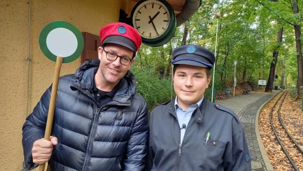 Ben und Anton stehen Seite an Seite. Anton ist Parkeisenbahner und hat einen entsprechenden blauen Hut auf. Ben hat einen ganz ähnlichen roten Hut auf und eine Anzeigekelle in der Hand. Beide werden vor dem Gleislauf der Bahn frontal fotografiert.
