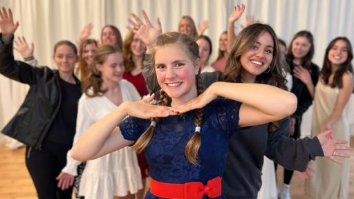 Jeanne steht vorn und hält ihre Fingerspitzen aneinander unter ihr Gesicht. Hinter ihr steht Sarah. Im Hintergrund sind weitere Schülerinnen der Musicalschule zu sehen.