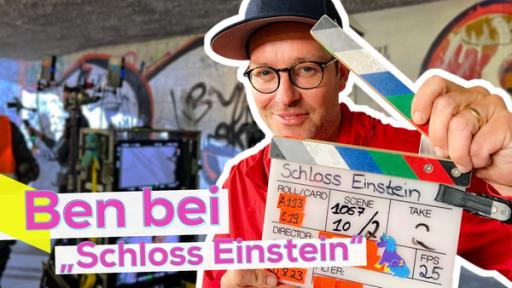 Rechte Bildhälfte Ben nah. Er hält eine geöffnete Filmklappe vor sich. Im Hintergrund eine Unterführung und Filmtechnik. Aufschrift: Ben bei Schloss Einstein.