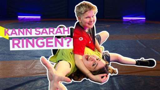 Sarah und Ringerin Josefine auf einer großen Matte. Beide sind fröhlich und imitieren einen Sieg über Sarah. Sarah liegt mit Rücken auf der Matte und streckt eine Hand Richtung Kamera. Josefine liegt über ihr. Aufschrift: Kann Sarah ringen?