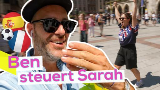 Links im Vordergrund Ben, der sich erschrocken, aber belustigt, die Hand vor den Mund hält. Links neben seinem Kopf ein Bild von einem Fußball und Flaggen von Spanien und Frankreich. Auf der rechten Seite Sarah, die jubelnd über einen öffentlichen Platz läuft. Aufschrift unten links: Ben steuert Sarah.