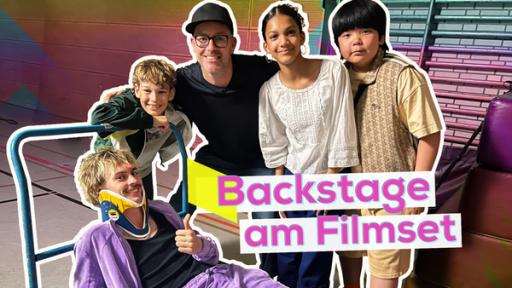 Noel Gabriel Kipp, Moderator Ben, Maïmouna Rudolph-Mbacké und Max Zheng eng nebeneinander. Davor liegt Cedric Eich mit Halskrause auf einen Rollwagen. Im Hintergrund eine Turnhalle. Aufschrift: Backstage am Filmset.